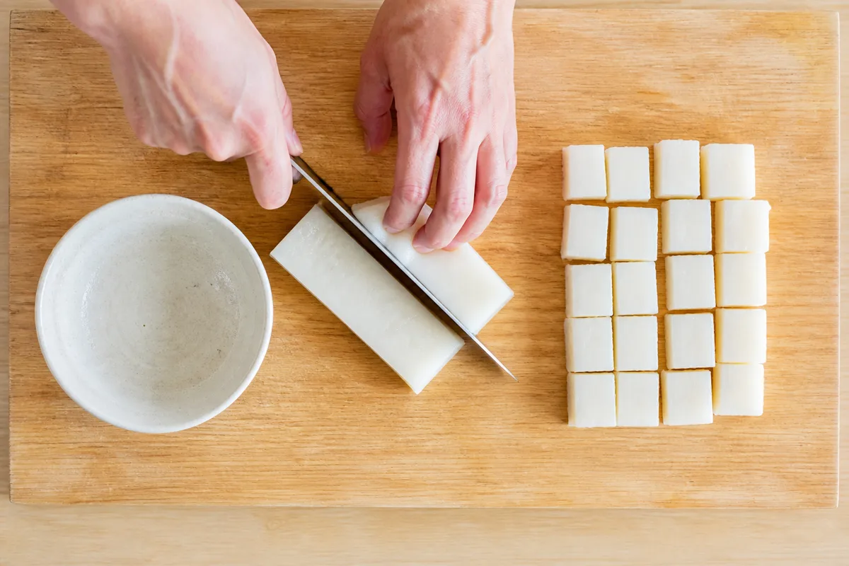 Mains découpant une galette de mochi en petits morceaux sur une planche de bois
